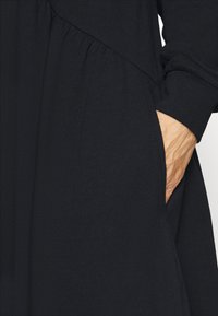 Svart klänning i tyg med en samlad midja och sidofickor; har långa ärmar och en slät yta.