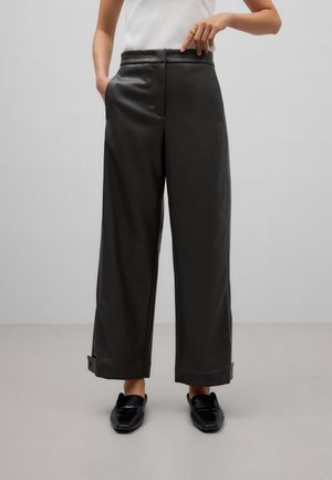 Personne portant un pantalon noir taille haute à jambes larges avec des ourlets retroussés et des chaussures noires sans lacets, pointant vers la taille.