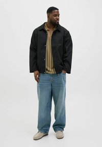 Svart nylonjacka med avslappnad passform över en beige randig skjorta, parat med lösa ljusblå jeans och beigea slip-on skor.