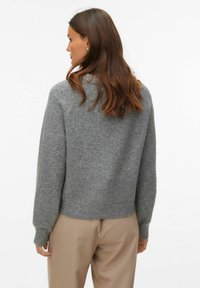 Szary wełniany sweter o luźnym kroju, z prążkowanymi mankietami i ściągaczem. Tył z okrągłym dekoltem i minimalistycznym wykończeniem. W zestawie z beżowymi spodniami.