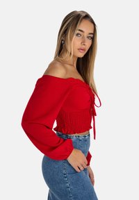 Rød off-shoulder bluse med rynket halsudskæring, tætsiddende smocket liv, og lange ærmer, parret med høj talje denimnederdel.