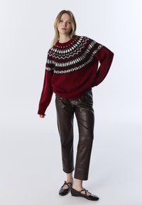 Pull en laine rouge avec un empiècement à motifs noir et blanc, associé à un pantalon en simili cuir marron et des ballerines noires avec des nœuds.