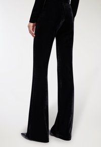 Czarne welurowe spodnie dzwony o gładkiej fakturze, z wysokim stanem i eleganckim krojem, przedstawione z tyłu. Widoczne stylowe buty na szpilce.