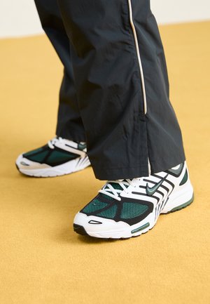 Czarne spodnie z beżowym paskiem po boku noszone z białymi, czarnymi i zielonymi butami Nike na żółtej podłodze.