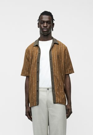 Homme portant une chemise à rayures marron à manches courtes par-dessus un T-shirt blanc et un pantalon gris clair, debout devant un fond uni.