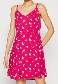 Abito senza maniche rosa con scollatura a V, caratterizzato da un motivo floreale bianco. Silhouette A-Line, tessuto leggero e tasche laterali.