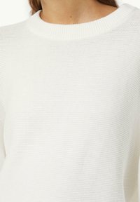 Vit stickad tröja med ribbad halsringning och strukturerad yta. Tyget verkar mjukt och lätt, lämpligt för vardagsbruk.