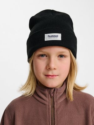 Dziecko z blond włosami do ramion nosi czarną czapkę Hummel Sportswear i brązową polarową kurtkę na zamek, stoi przodem.