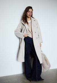 Beige uldblandingsfrakke med brede revers og dobbelte knapper, parret med en lys sweater og mørke denim flared bukser. Grå pels skuldertaske.