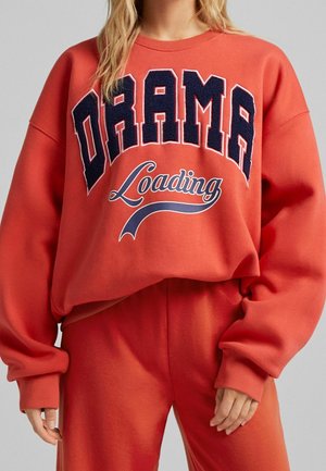 Vrouw draagt een oversized rode trui en broek, op de trui staat in grote marineblauwe en witte letters 'DRAMA Loading'.