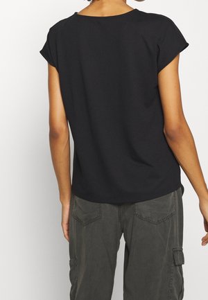 Femme portant un haut noir à manches courtes et un pantalon cargo gris foncé vue de dos sur un fond uni.