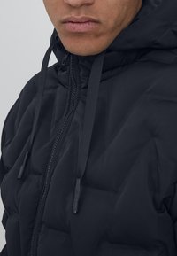 Svart pufferjakke med hette, med glidelås og snøring. Materialet virker mykt med en quiltet tekstur og subtil glans.