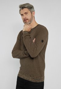 Brązowy sweter z dzianiny o teksturowanym wzorze, ze ściągaczami na rękawach i okrągłym dekoltem. Zawiera małe czarne logo na lewym rękawie.