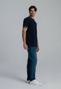 Granatowa koszulka polo z kontrastowym kołnierzykiem i wykończeniem rękawów, zestawiona z niebieskimi dżinsami oraz białymi trampkami. Model pozuje bokiem na neutralnym tle.