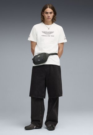 Junge Person mit schulterlangem Haar, trägt ein weißes T-Shirt des Aston Martin Formel-1-Teams, schwarze geschichtete Shorts und Hose, schwarze Bauchtasche.