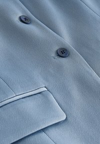 Hele sinine jakk siledast kangast, millel on kaks tumedat nööpi ja korrapäraselt vormitud krae ning väike tasku detail.