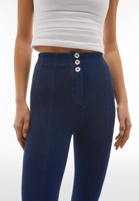 Jeans blu a vita alta con un leggero luccichio, dotati di tre bottoni bianchi nella parte frontale, un tessuto liscio e una vestibilità aderente.