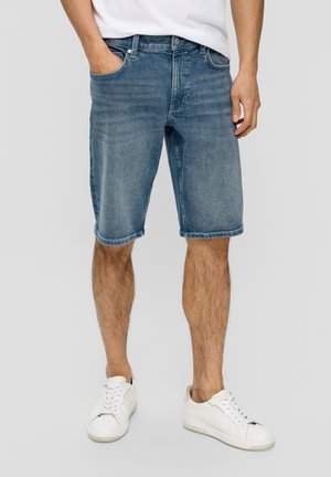 Denimshorts i en falmet blå vask med fem lommer og en lige pasform, med opviklede kanter. Kombineret med hvide sneakers.