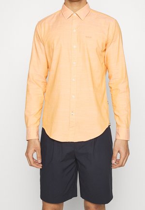 Hombre vistiendo una camisa de botones de manga larga color durazno y pantalones cortos negros hasta la rodilla, de pie frente a un fondo claro y liso.