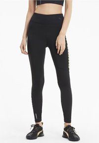 Czarne legginsy sportowe z wysokim stanem, z teksturowanymi akcentami i złotym logo po boku, w zestawie z czarnymi sneakersami.