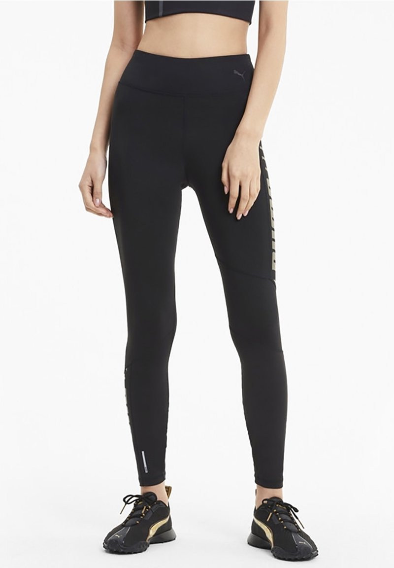 Czarne legginsy sportowe z wysokim stanem, z teksturowanymi akcentami i złotym logo po boku, w zestawie z czarnymi sneakersami.