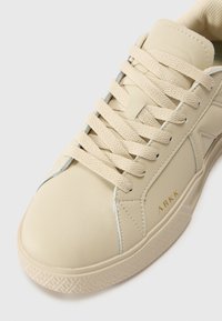 Beige leren sneaker met platte beige veters, textuurzool, minimalistisch ontwerp en logo detail in goud aan de zijkant.