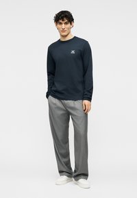 Νέος άνδρας που φοράει σκούρο μακρυμάνικο πουκάμισο, ανοιχτό γκρι παντελόνι και άσπρα αθλητικά παπούτσια, στέκεται με το ένα χέρι στην τσέπη.