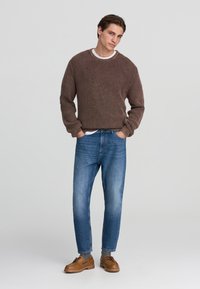 Maglione a coste marrone con maniche lunghe, abbinato a jeans di denim a sigaretta blu chiaro e scarpe marroni stringate. Canottiera bianca semplice visibile.