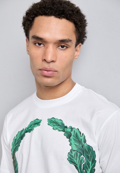 White cotton t-shirt featuring a green leaf graphic design. Short sleeves, round neckline, and smooth texture.