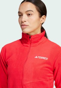 Rote Fleecejacke mit hohem Kragen, ausgestattet mit einem Reißverschluss und dem weißen "TERREX"-Logo auf der linken Seite. Weicher Stoff und taillierte Passform.