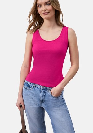 Kobieta nosząca jaskraworóżowy top bez rękawów i jasnoniebieskie dżinsy, trzyma brązową torebkę, jedną ręką w kieszeni.