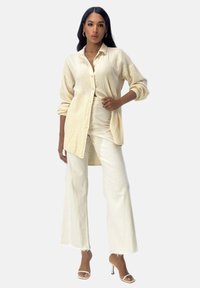 Camicia con bottoni in tessuto crema texturizzato, abbinata a pantaloni a gamba larga crema. L'outfit presenta una vestibilità rilassata e una leggera lucentezza.