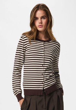 Jeune femme portant un cardigan boutonné à rayures horizontales marron et blanc avec un pantalon marron foncé, debout sur un fond blanc.