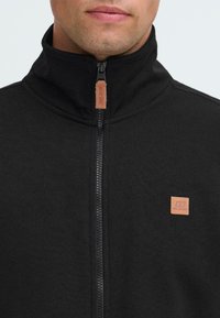 Mann iført en svart zip-up jakke med høy krage og lærmerker på glidelås og bryst.