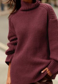 Person, der einen grob gestrickten, weinroten Rollkragenpullover mit hochgeschobenen Ärmeln trägt, deren Hand in einer Tasche steckt.