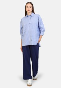Camicia button-up a righe blu e bianche con colletto, maniche lunghe e vestibilità rilassata, abbinata a pantaloni wide-leg blu navy e sneakers bianche.