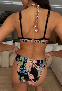 Setul de bikini prezintă un model floral în culori vii pe un fundal negru, bretele reglabile și o închizătoare din metal auriu la spate.