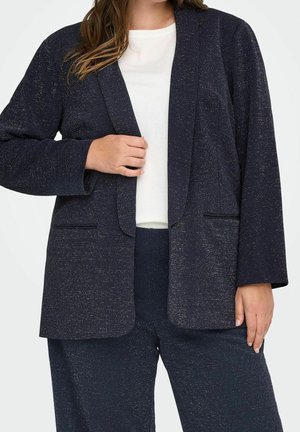 Kvinde iført en mørk plettet blazer og matchende bukser over en hvid top, mens hun holder blazeren med den ene hånd.