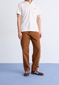 Bílý polo trikotu s krátkým rukávem a v-výstřihem, doplněný červeným logem, v kombinaci s hnědými bavlněnými kalhotami a burgundskými teniskami s bílými pruhy.