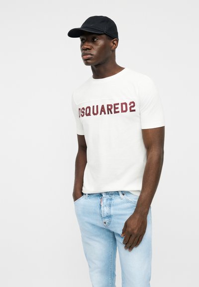 Homme portant une casquette noire, un t-shirt blanc Dsquared2 et un jean bleu clair, debout les mains dans les poches.