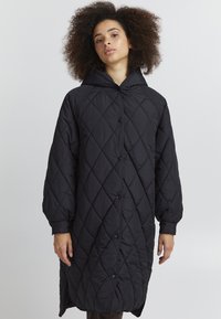Manteau long matelassé noir avec capuche, présentant un motif en losanges, des boutons sur le devant et des manches larges. Texture lisse et coupe oversize.