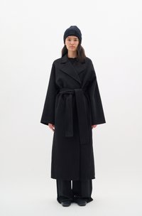 Black wool coat with wide sleeves, belted waist, and lapel collar; paired with a dark ribbed knit beanie and black pants.