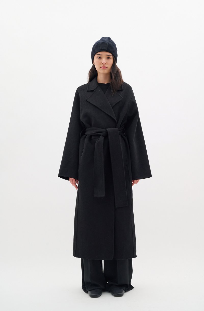 Black wool coat with wide sleeves, belted waist, and lapel collar; paired with a dark ribbed knit beanie and black pants.