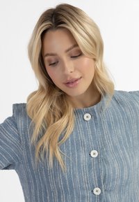 Lichtblauwe, gestructureerde blouse met verticale witte strepen, voorzien van een ronde halslijn en vijf witte knopen aan de voorkant.