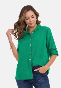 Groene button-up shirt met twee borstzakken, opgerolde mouwen en houten knopen. Gestructureerde stof met een casual pasvorm. Gepaard met donkere jeans.
