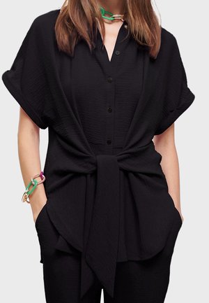 Chemise noire à manches courtes, coupe ample, boutonnée à l'avant, avec un détail de taille nouée. Tissu texturé. Le mannequin porte des bracelets colorés.