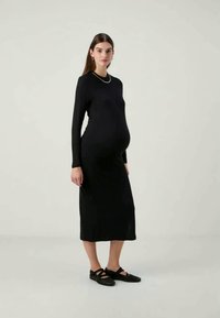 Μαύρο φόρεμα με μακριά μανίκια, στενή σιλουέτα, κοπή μέχρι το γόνατο, ραβδωτή υφή και στρογγυλή λαιμόκοψη, σε συνδυασμό με μαύρα φλατ παπούτσια.