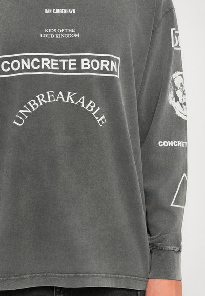 Suure plaanis tumehall pikka varrukaga särk, millel valged tekstid: "HAN KJØBENHAVN," "KIDS OF THE LOUD KINGDOM," "CONCRETE BORN," ja "UNBREAKABLE."
