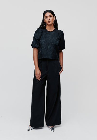 Čierny zamatový crop top s nafúknutými rukávmi, skombinovaný s čiernymi nohavicami s vysokým pásom a striebornými topánkami s ostrou špičkou. Hladká štruktúra, elegantný silueta.
