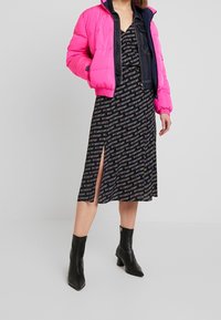 Žena v růžové péřové bundě přes černé midi šaty s vzorem a bočním rozparkem, černých lodičkách s špičatou špičkou a džínové bundě pod tím.